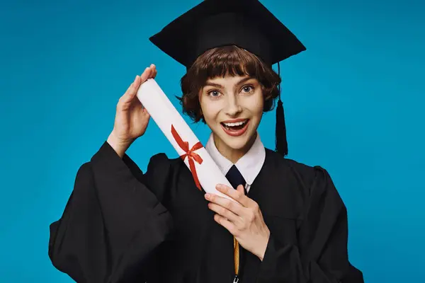 Allegra ragazza del college in abito e cappello accademico che tiene il suo diploma con orgoglio su blu, laurea — Foto stock