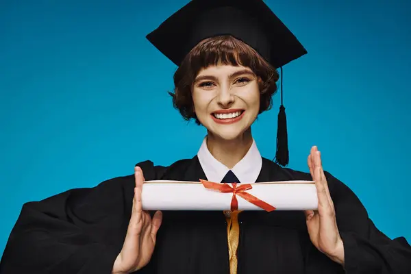 Positive grad college girl in gown and academic cap holding her diploma with pride on blue — Stock Photo