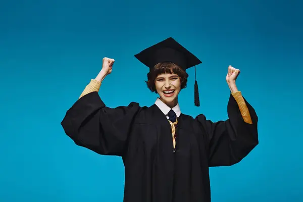Eccitato laureato ragazza del college in abito e cap alzando i pugni su sfondo blu, realizzazione — Foto stock