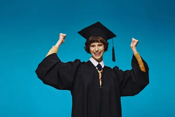 Excitado graduado chica universitaria en vestido y la tapa levantando puños sobre fondo azul, logro - foto de stock