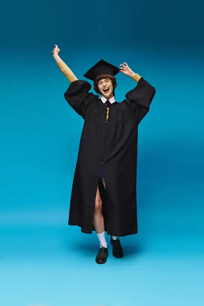 Eccitato laureato ragazza del college in abito e cap alzando i pugni su sfondo blu, successo — Foto stock