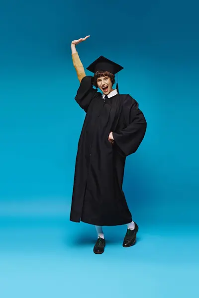 Animado graduado menina da faculdade em vestido e capacete levantando e gritando de alegria no fundo azul — Fotografia de Stock