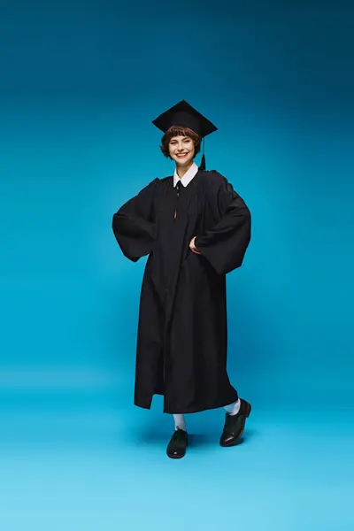 Fröhliche Hochschulabsolventin in Kleid und Mütze lächelnd auf blauem Hintergrund, Errungenschaft — Stockfoto