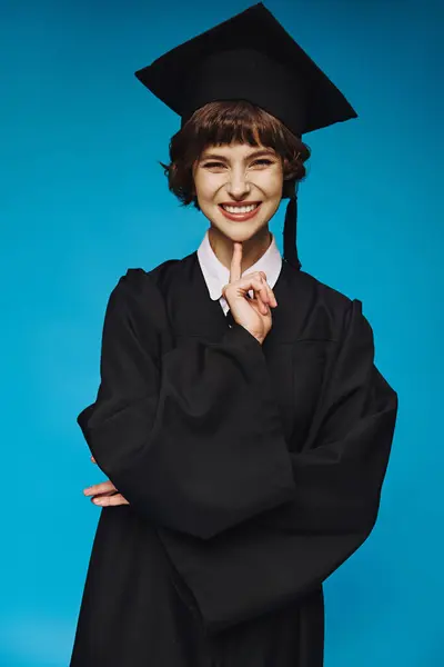 Положительный выпускник колледжа девушка в платье и кепке улыбаясь на синем фоне, достижение — стоковое фото