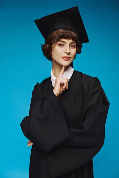 Young graduated college girl in gown and cap standing on blue background, accomplishment — Stock Photo
