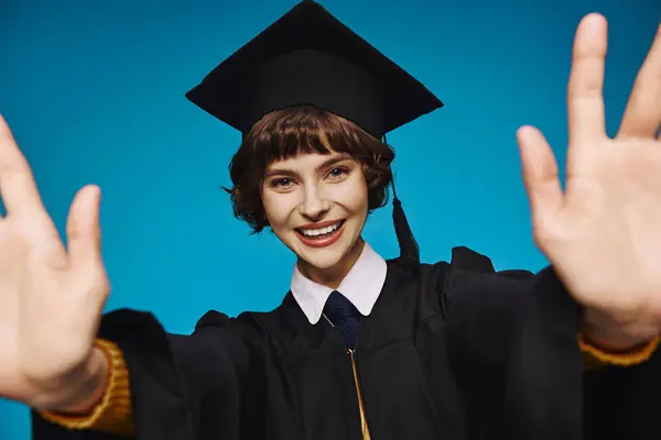 Happy graduate college girl in academic cap with outstretched hands on blue backdrop, accomplishment — Stock Photo