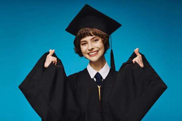 Alegre graduado chica universitaria en la tapa académica haciendo signo de corazón con los dedos en azul telón de fondo - foto de stock