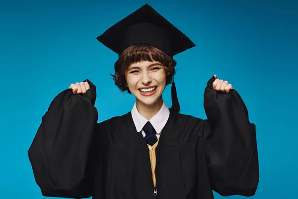 Young brunette graduate college girl in academic cap feeling happy on blue background, success — Stock Photo