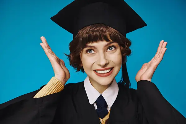 Jovem e alegre menina da faculdade de pós-graduação em cap acadêmico gesticulando com as mãos, fundo azul — Fotografia de Stock