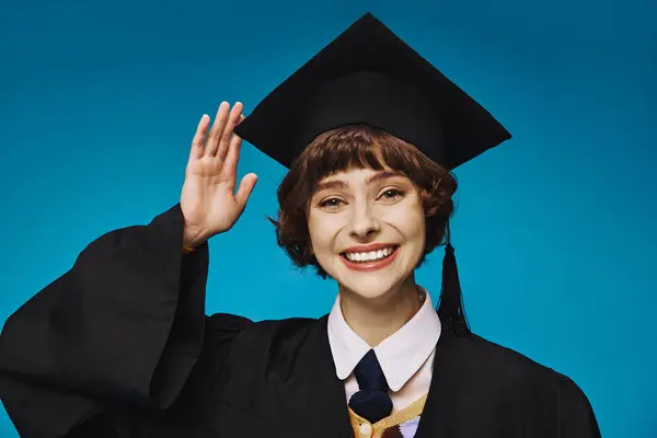 Young and cheerful graduate college girl in academic cap saluting with hand, blue background — Stock Photo