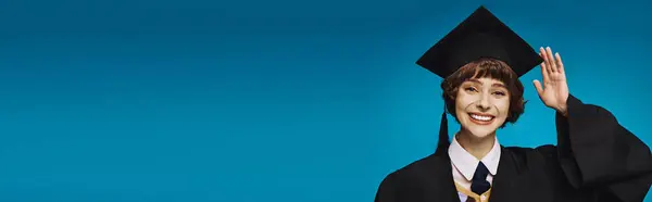 Young and cheerful graduate college girl in academic cap saluting with hand, blue background banner — Stock Photo