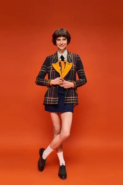 Full length of stylish student in checkered blazer and skirt on orange background, college uniform — Stock Photo