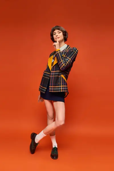 Stylish student in checkered blazer and skirt showing hush on orange background, college uniform — Stock Photo