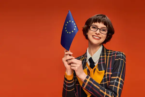 Happy young college girl in uniform and glasses holding EU flag on vibrant orange background — Stock Photo