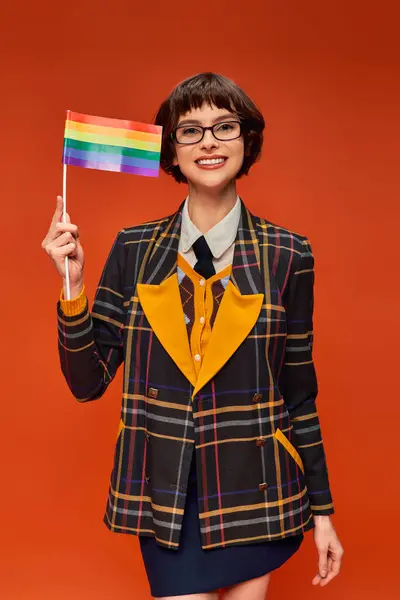 Радісна молода дівчина коледжу в уніформі і окулярах тримає прапор ЛГБТ і стоїть на помаранчевому фоні — стокове фото