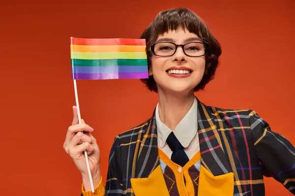 Positivo giovane ragazza del college in uniforme e occhiali con bandiera lgbt su sfondo arancione, orgoglio — Foto stock