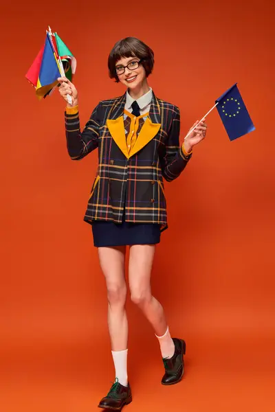 Happy young college girl in uniform and glasses holding EU and different flags on orange background — Stock Photo
