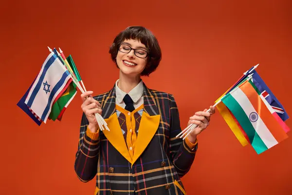 Menina da faculdade feliz em seu uniforme e óculos segurando várias bandeiras e de pé em pano de fundo laranja — Fotografia de Stock
