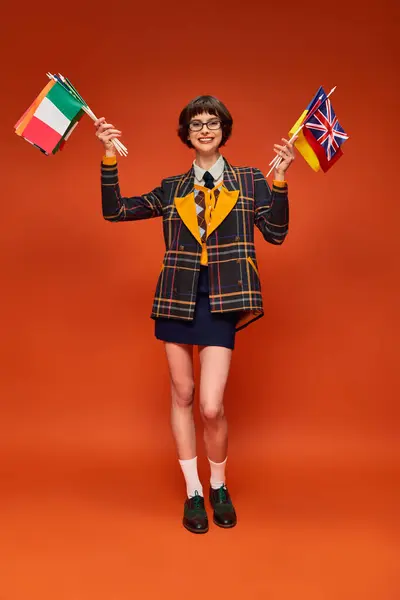 Happy student girl in her uniform and glasses holding variety of flags on orange background — Stock Photo