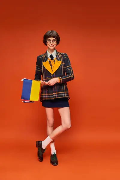 Happy young college girl in uniform and glasses holding Ukrainian flag on orange background — Stock Photo