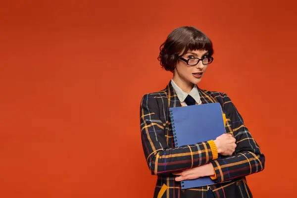 Estudiante reflexivo en uniforme universitario y gafas que sostienen el cuaderno sobre fondo naranja, chica inteligente - foto de stock
