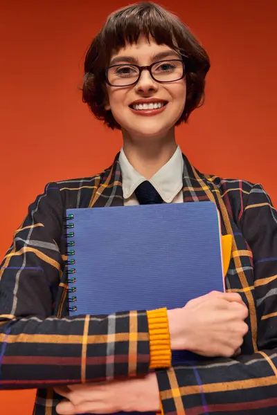 Étudiant heureux en uniforme à carreaux et lunettes tenant un carnet sur fond orange, fille de l'université — Photo de stock