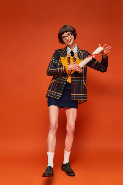 Excitada chica universitaria graduada en uniforme y gafas sosteniendo su diploma en vibrante fondo naranja - foto de stock