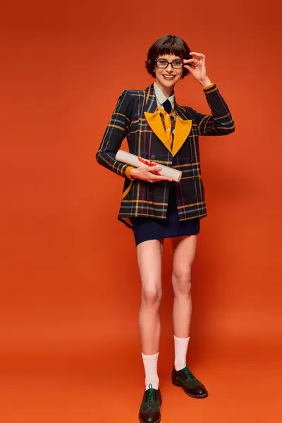 Alegre graduado chica universitaria en uniforme y gafas celebración de su diploma en vibrante fondo naranja - foto de stock