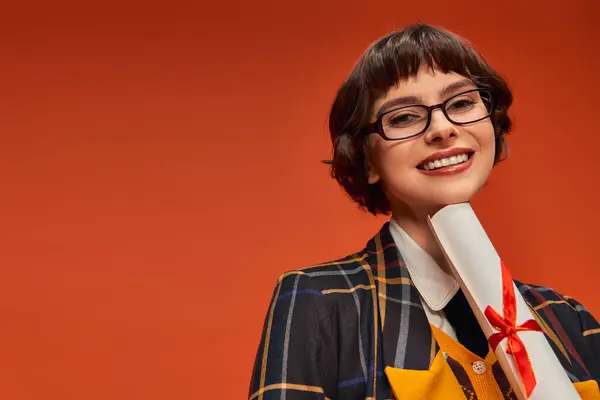 Positive college girl in uniform and glasses holding her graduation diploma on orange backdrop — Stock Photo