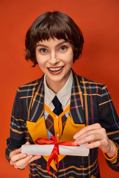 Ritratto di eccitata ragazza del college in uniforme a scacchi possesso diploma di laurea su sfondo arancione — Foto stock