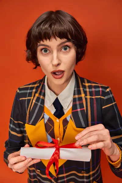 Portrait of shocked college girl in checkered uniform holding graduation diploma on orange backdrop — Stock Photo