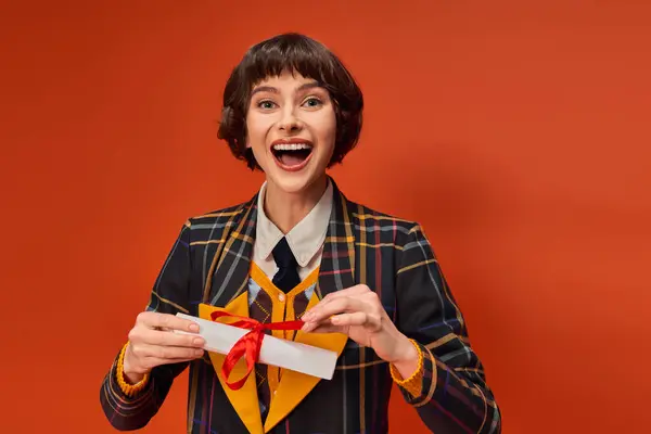 Porträt eines aufgeregten College-Mädchens in karierter Uniform, das sein Diplom vor orangefarbenem Hintergrund hält — Stockfoto
