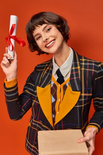 Ritratto di eccitata ragazza del college in uniforme a scacchi in possesso di libri e diploma su sfondo arancione — Foto stock