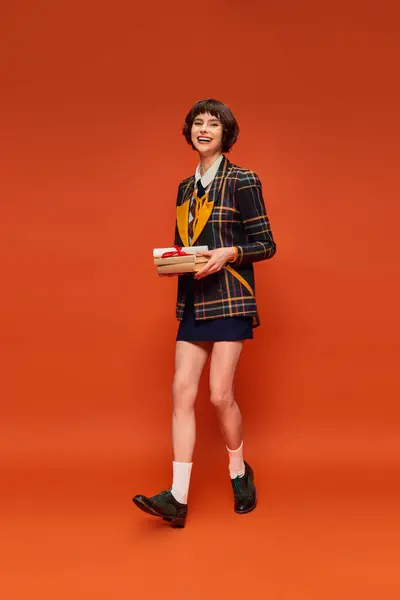 Retrato de estudiante positivo en uniforme universitario sosteniendo libros y diploma sobre fondo naranja - foto de stock