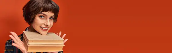 Banner of smiling college girl in uniform holding books near chin on orange background, knowledge — Stock Photo