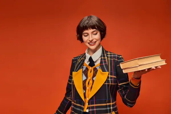 Happy college girl in uniform holding stack of books in hand on orange background, knowledge — Stock Photo