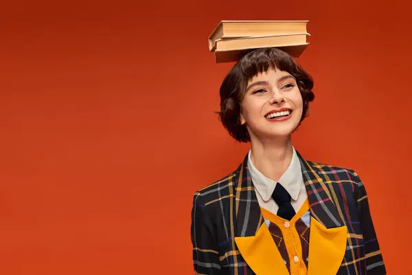 Optimistic college girl in uniform holding stack of books on hand on orange background — Stock Photo