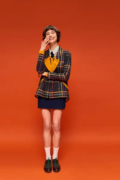 Chica universitaria optimista en uniforme a cuadros sonriendo sobre fondo naranja, feliz vida estudiantil - foto de stock