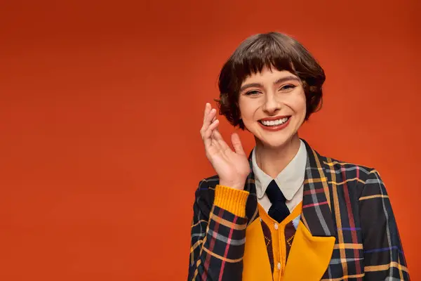 Optimiste collège fille à carreaux uniforme agitant la main sur fond orange, heureuse vie étudiante — Photo de stock