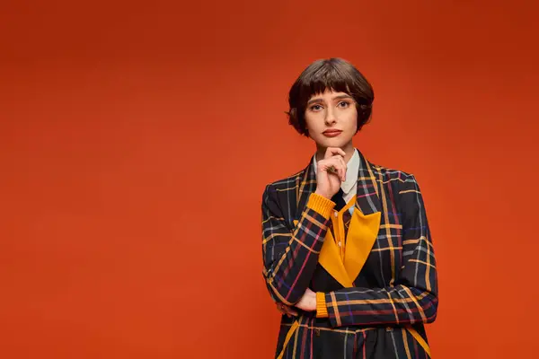 Riflessivo college girl con i capelli corti posa in uniforme a scacchi su sfondo arancione, decisione — Foto stock