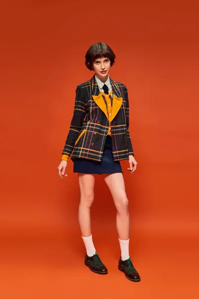 Full length of college girl in checkered blazer and footwear with socks standing on orange backdrop — Stock Photo