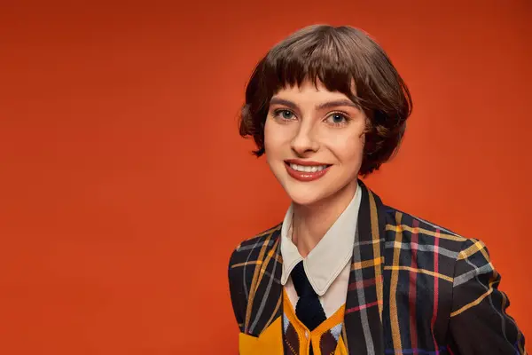 Chica estudiante optimista y joven en uniforme a cuadros de la universidad sonriendo sobre fondo naranja - foto de stock