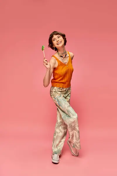 Felice e divertente giovane donna in abiti vivaci tenendo il suo lecca-lecca su sfondo rosa, amante delle caramelle — Foto stock