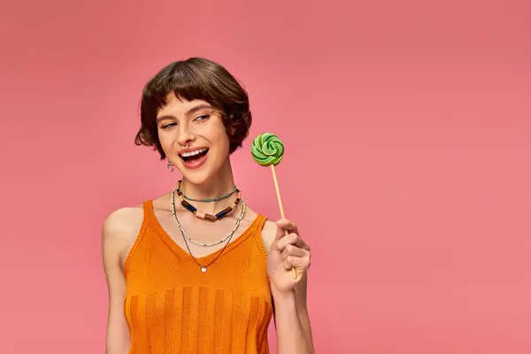 Cheerful young woman in orange knitted tank top holding colorful sweet lollipop on pink backdrop — Stock Photo