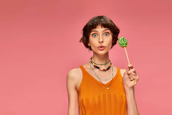 Amazed young woman in orange knitted tank top holding colorful sweet lollipop on pink backdrop — Stock Photo