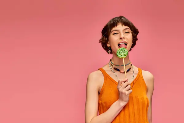 Happy young woman in orange knitted tank top licking colorful sweet lollipop on pink backdrop — Stock Photo
