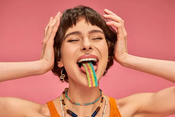 Excité jeune femme dans sa vingtaine dégustation sucré et aigre bande de bonbons sur sa langue, fond rose — Photo de stock