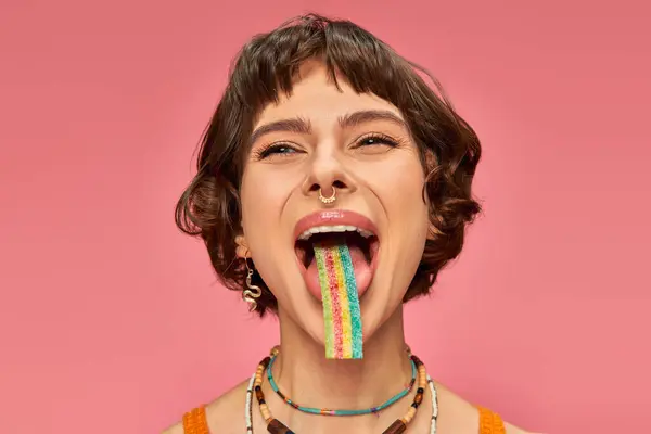 Joyeuse jeune femme dans la vingtaine dégustation sucrée et aigre bande de bonbons sur sa langue, fond rose — Photo de stock