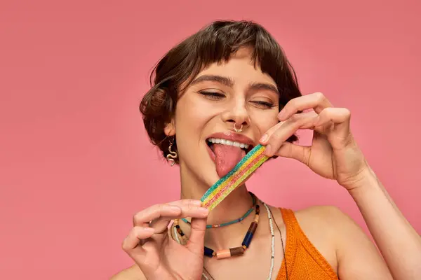 Joyeuse jeune femme dans sa vingtaine lécher sucré et aigre bande de bonbons sur sa langue, fond rose — Photo de stock