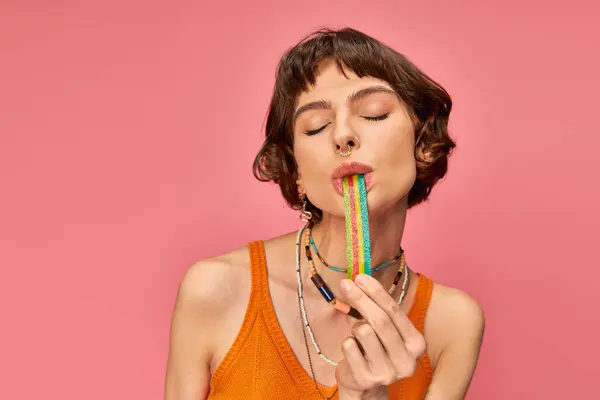 Portrait of cheerful young woman in her 20s biting sweet and sour candy strip on pink background — Stock Photo